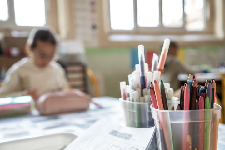 une enfant qui écrit ou dessine en arrière plan, au premier plan des crayons et feutres